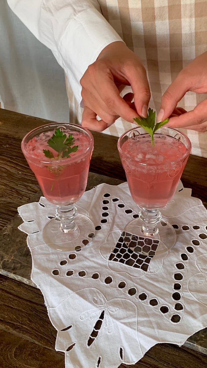 Aromatic pink cocktails with garnish served on a lace cloth atop a wooden table.